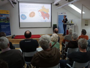 Blick von hinten in einen Vortragsraum. Zu sehen sind die Rücken der Zuhörenden, die auf die Referentin bzw. ihre Präsentation blicken.