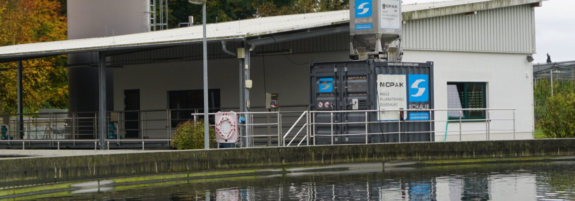 Blick auf die mobile Pulveraktivkohledosieranlage MOPAK in einem Container zwischen einem Klärbecken und einem Gebäude.