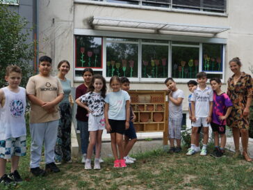 Blick auf eine Gruppe von Dritt- und Viertklässlern, die links und rechts neben einem Bienenhotel stehen und in die Kamera schauen.